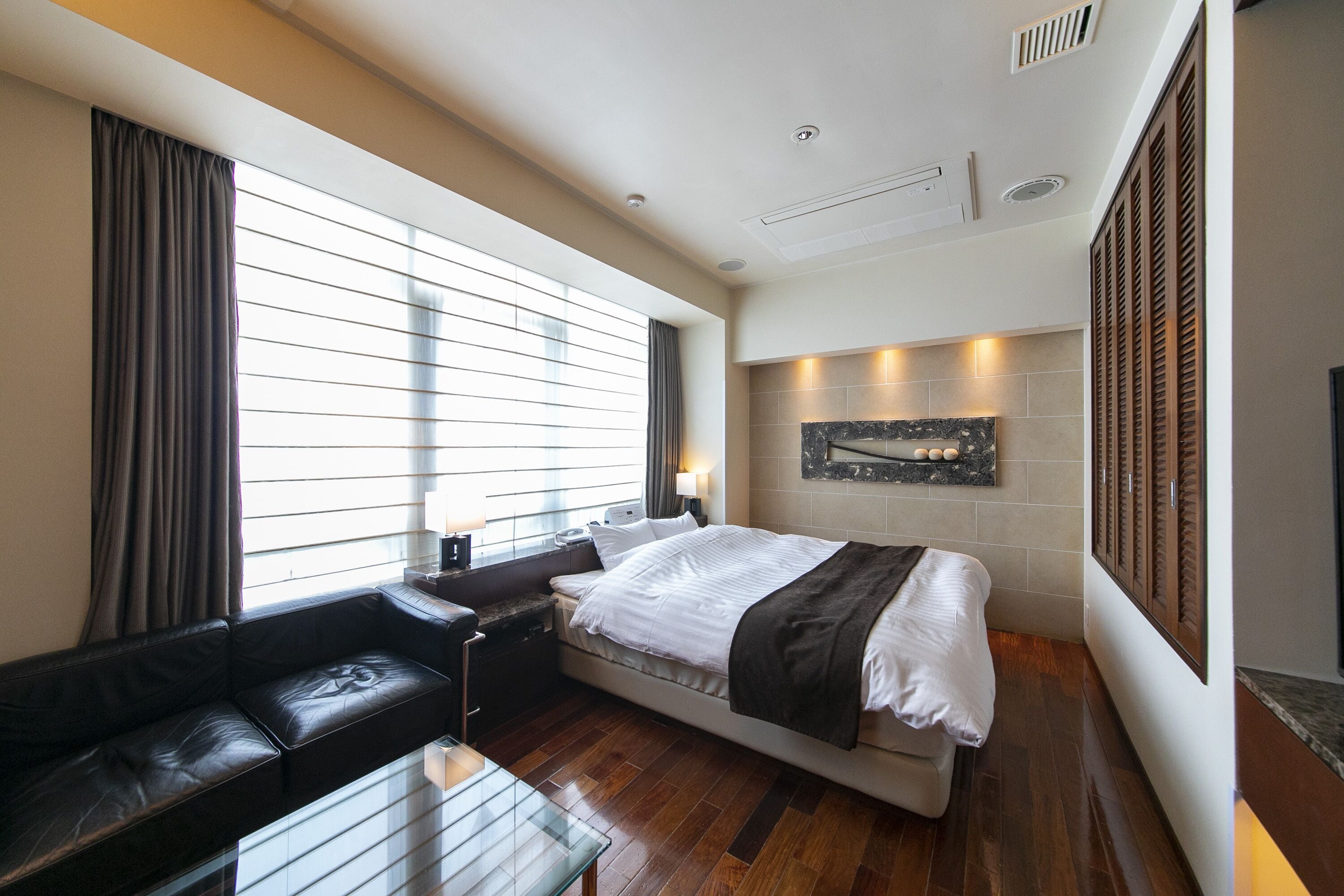 A modern hotel room with a large bed, a sofa, a glass coffee table, and a view of the city through large windows.
