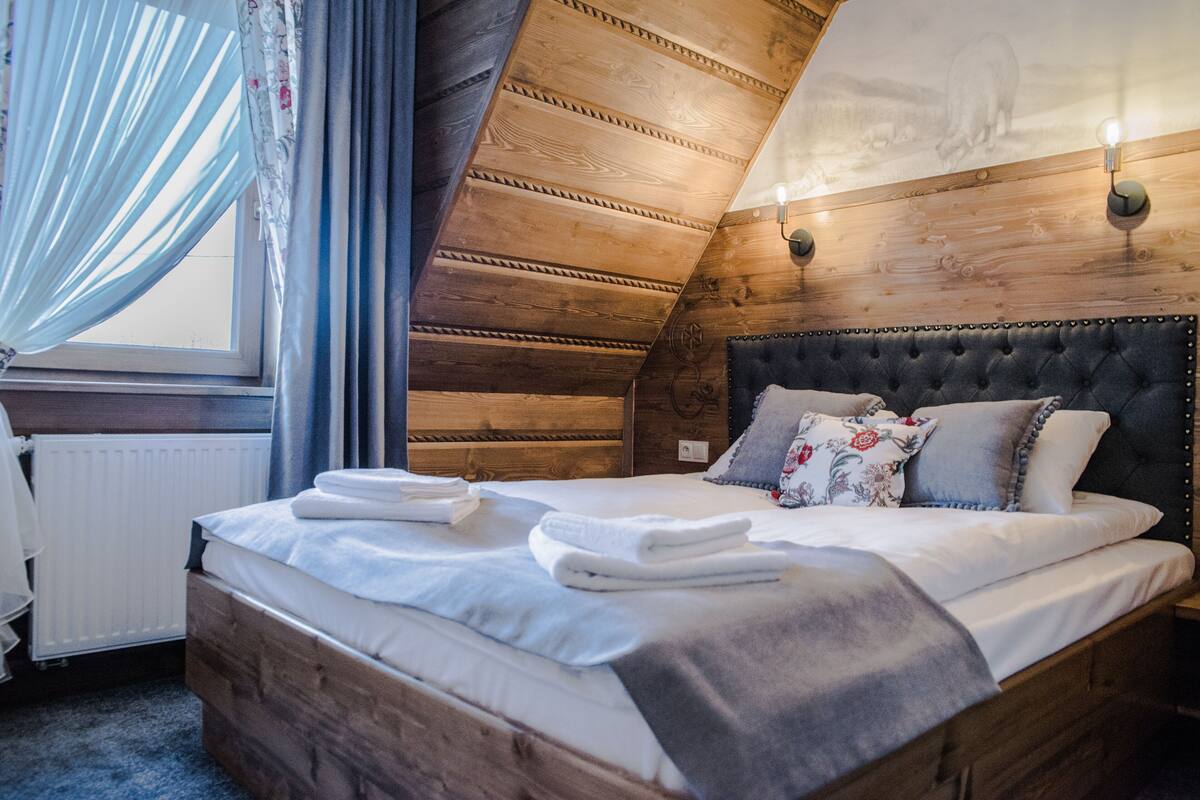 A bedroom with a wooden ceiling, a bed with white linens, and a window with curtains.