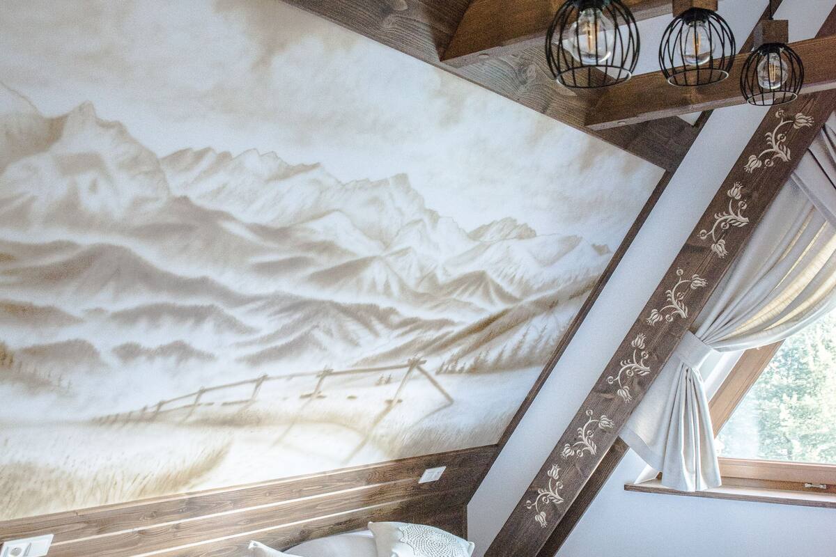 A bedroom with a wooden ceiling, a bed with white linens, and a mountain landscape mural on the wall.