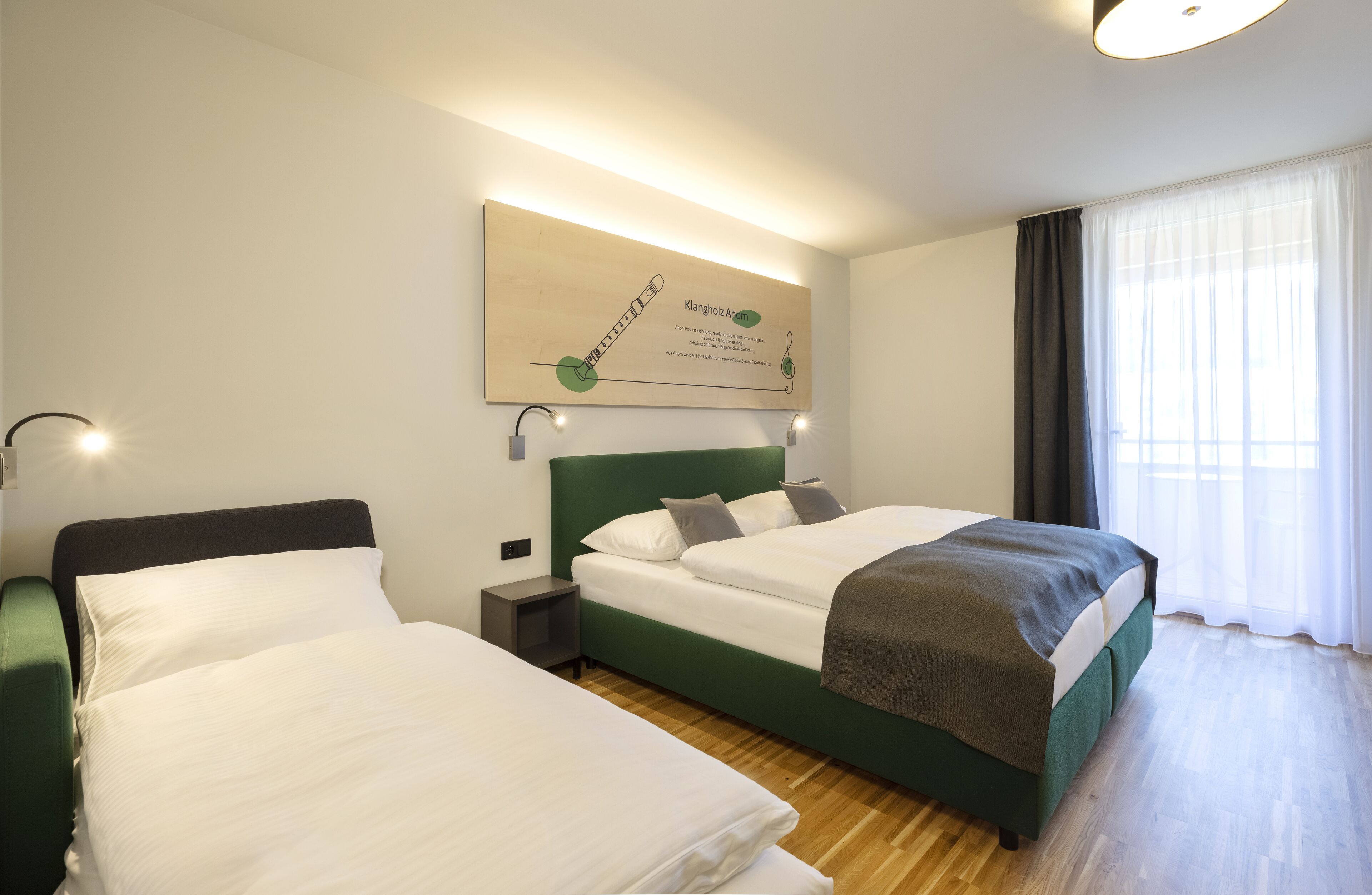 A hotel room with two beds, a wooden floor, a large window with curtains, and a wall-mounted artwork.