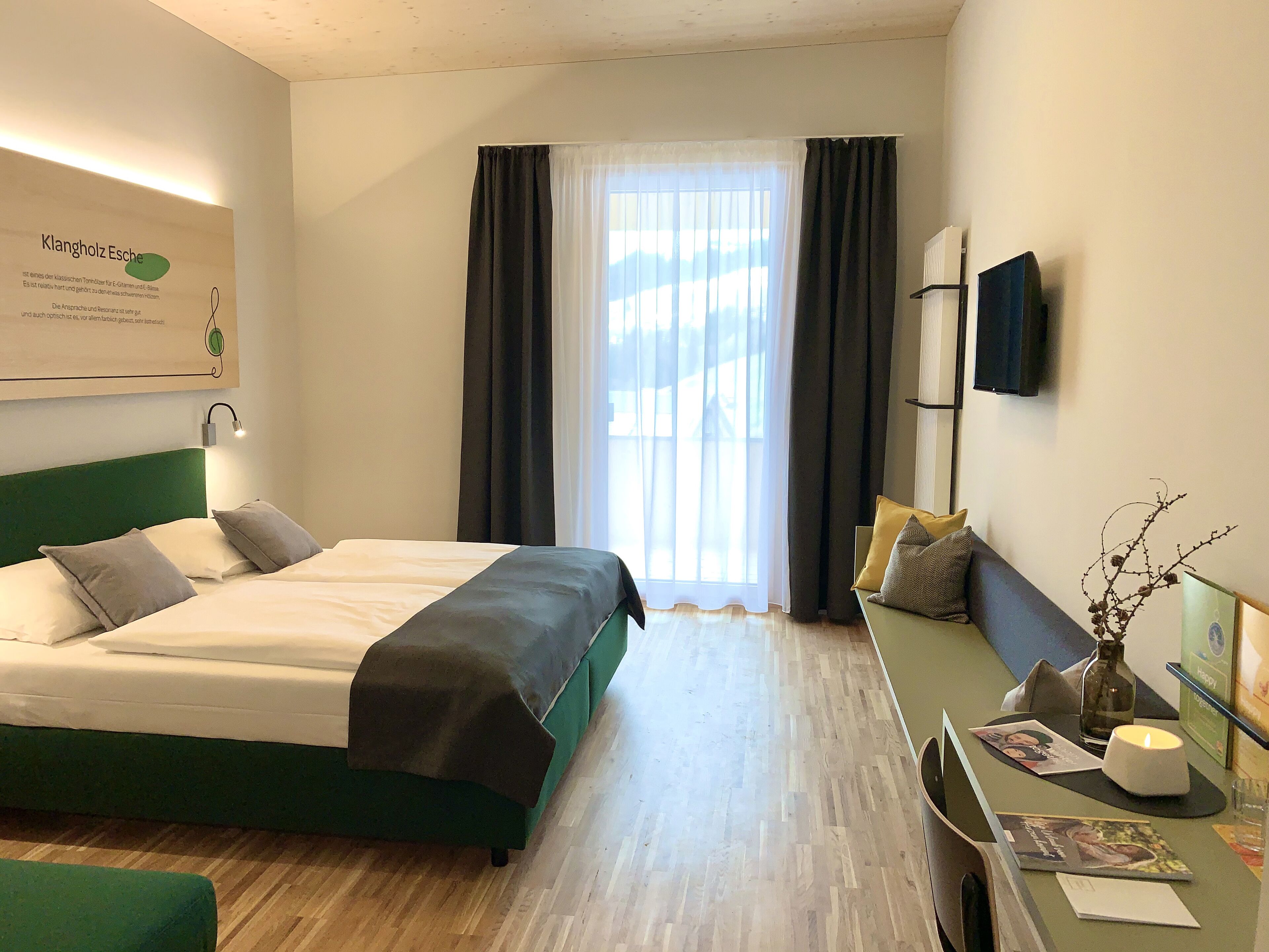 A hotel room with a bed, a desk with a chair, a TV mounted on the wall, and a window with curtains.