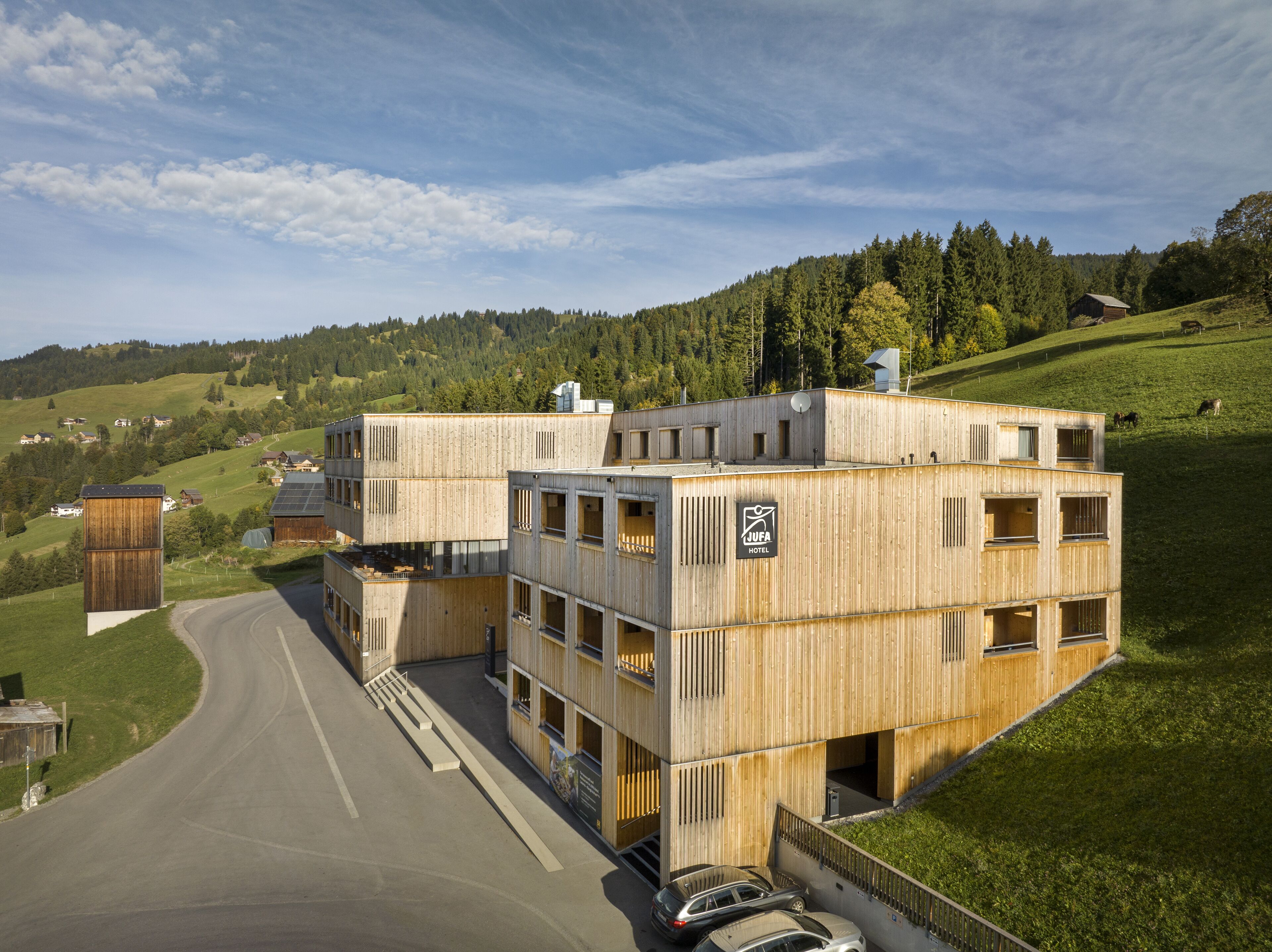A modern wooden hotel with multiple floors and balconies, located on a hillside with a view of the surrounding landscape.