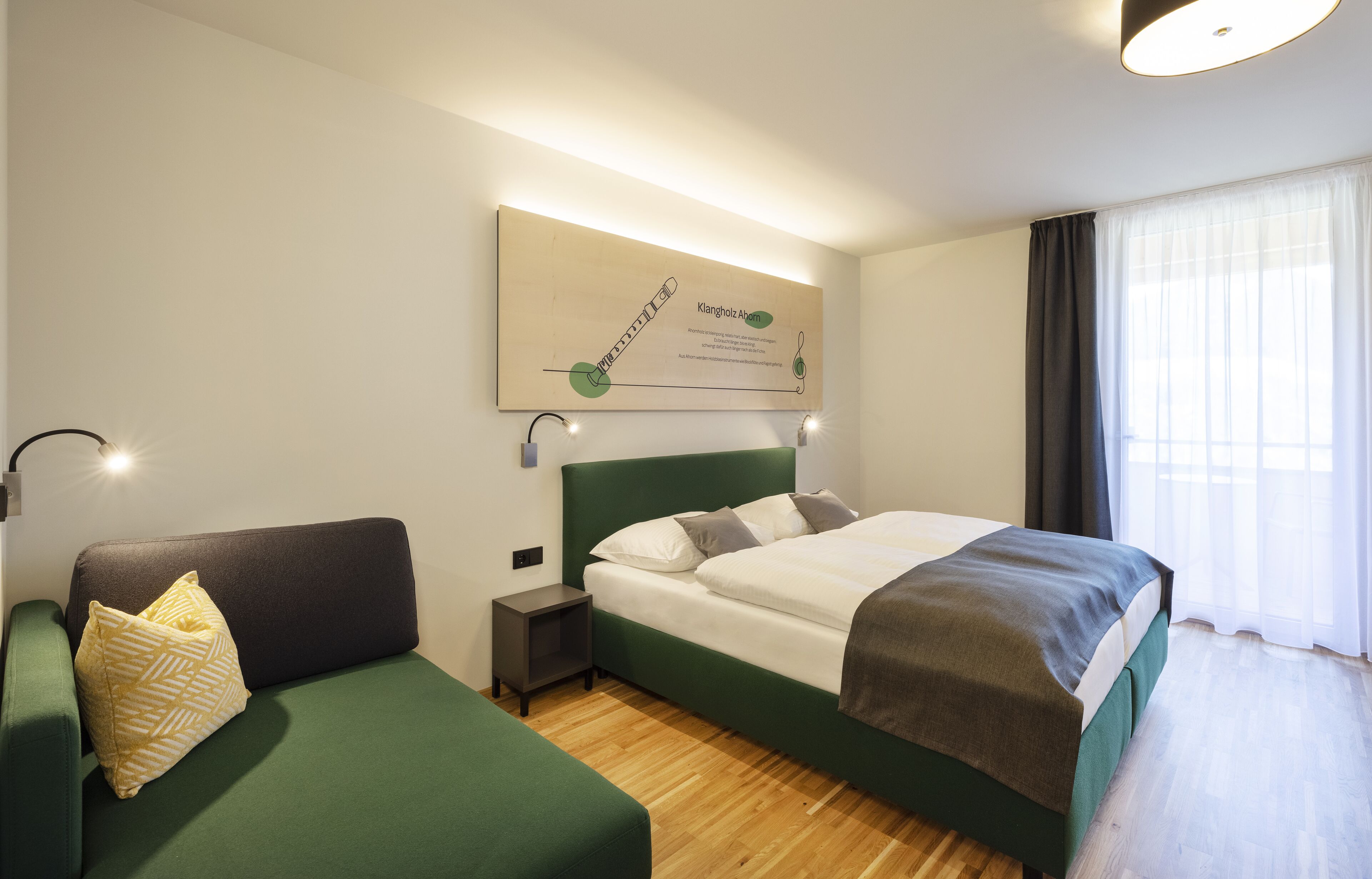 Habitació d'hotel moderna amb un sofà verd, un llit ben tendit, una petita taula de nit i una finestra gran amb cortines.