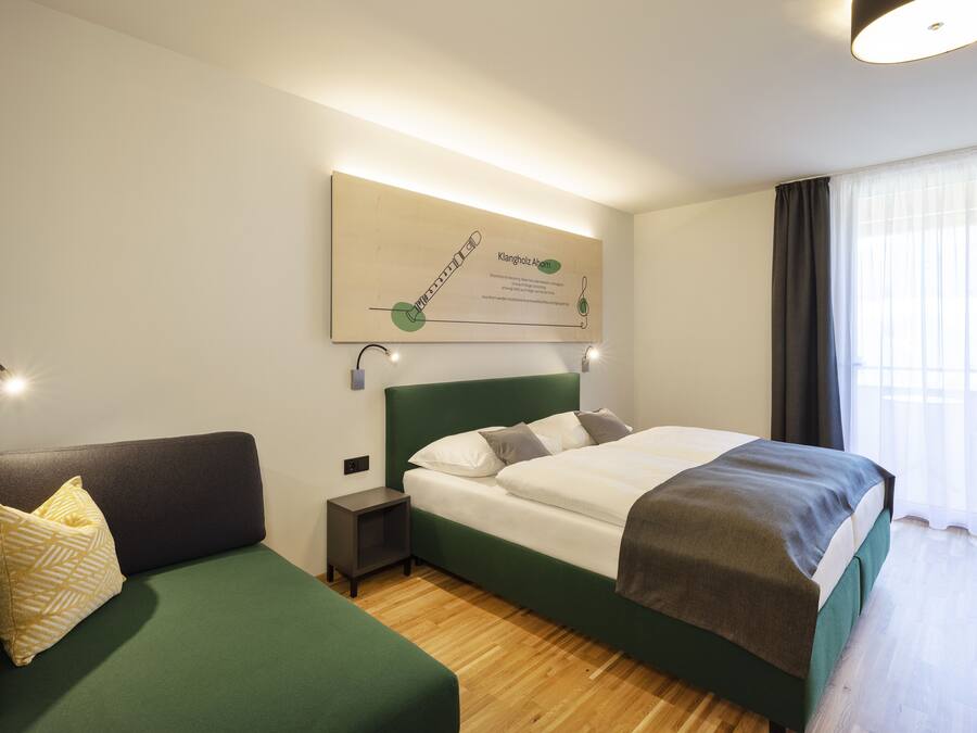 Habitació d'hotel moderna amb un sofà verd, un llit ben tendit, una petita taula de nit i una finestra gran amb cortines.