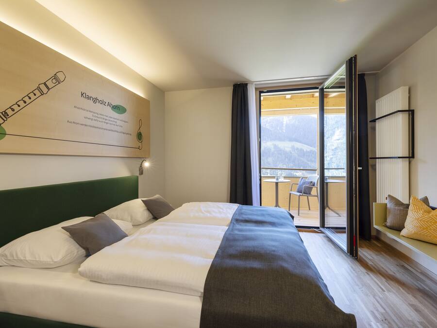 A hotel room with a bed, a view of mountains, a balcony, and a wall-mounted information board.