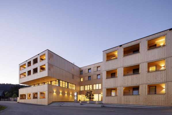 A modern multi-story building with a wooden facade, multiple balconies, and illuminated windows. - JUFA Hotel Laterns - Klangholzhus (Laterns)
