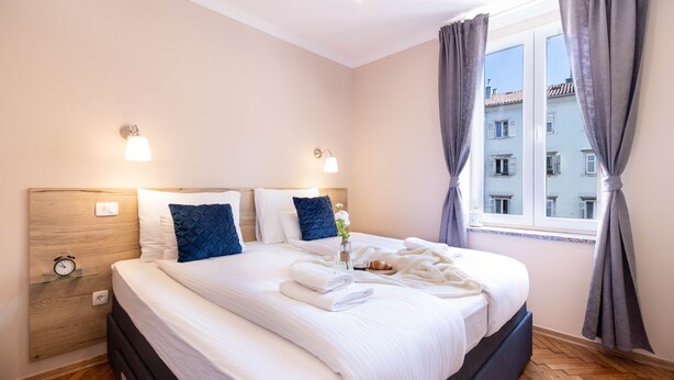 Un lit bien fait, avec du linge de lit blanc et des coussins bleus, une table de chevet avec une lampe et une fenêtre avec des rideaux.