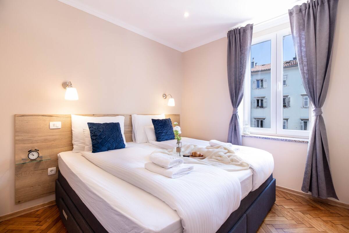 Una cama bien hecha con sábanas blancas y almohadas azules, una mesita de noche con lámpara, y una ventana con cortinas.