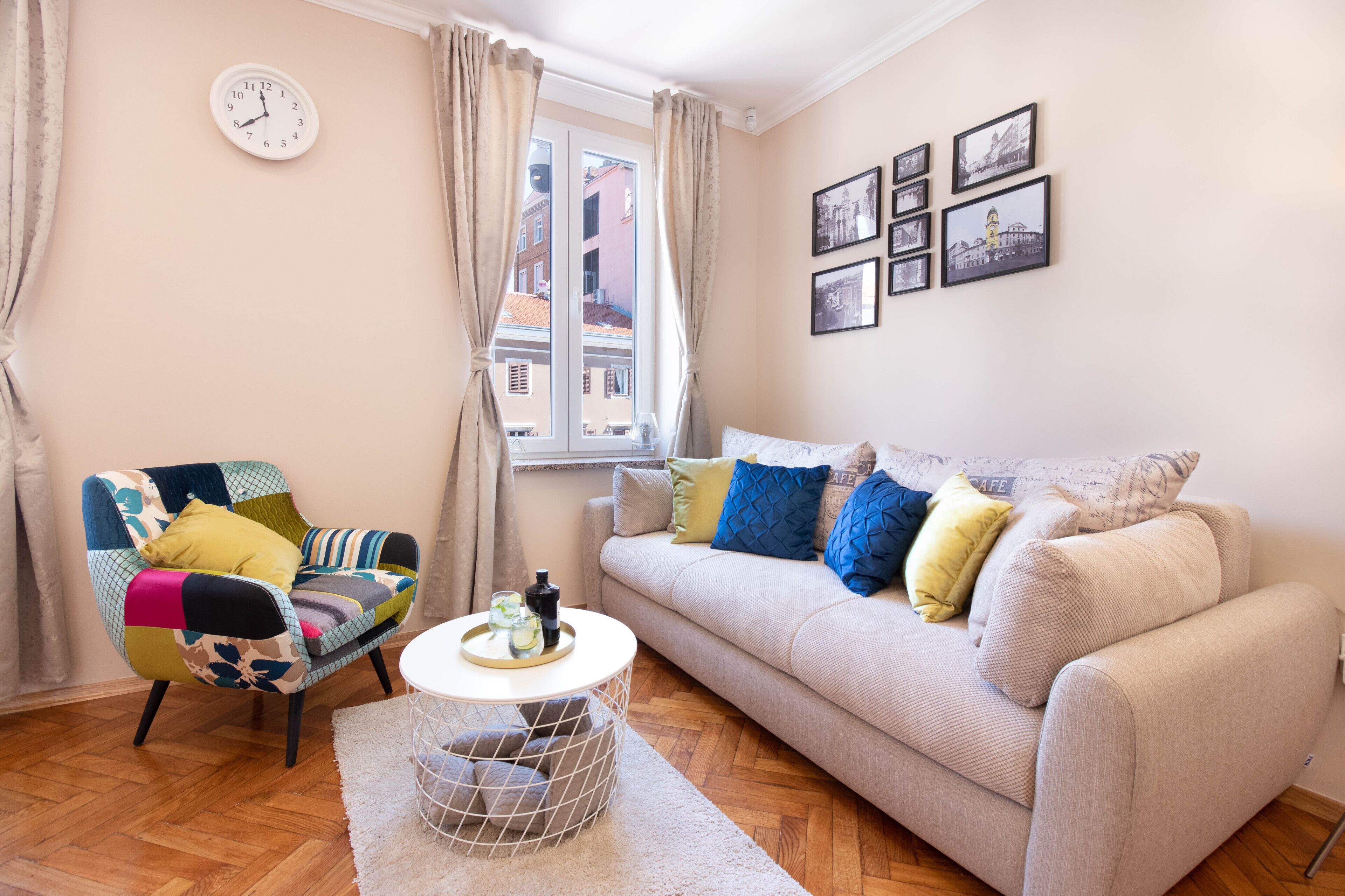 Ein gemütliches Wohnzimmer mit einem farbenfrohen Sessel, einer Couch mit gemusterten Kissen, einem runden Couchtisch und einem Fenster mit Vorhängen.