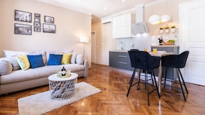 Un salon moderne avec un canapé, une table basse et un coin repas avec des chaises. On aperçoit au fond une cuisine équipée de placards et d’un évier.