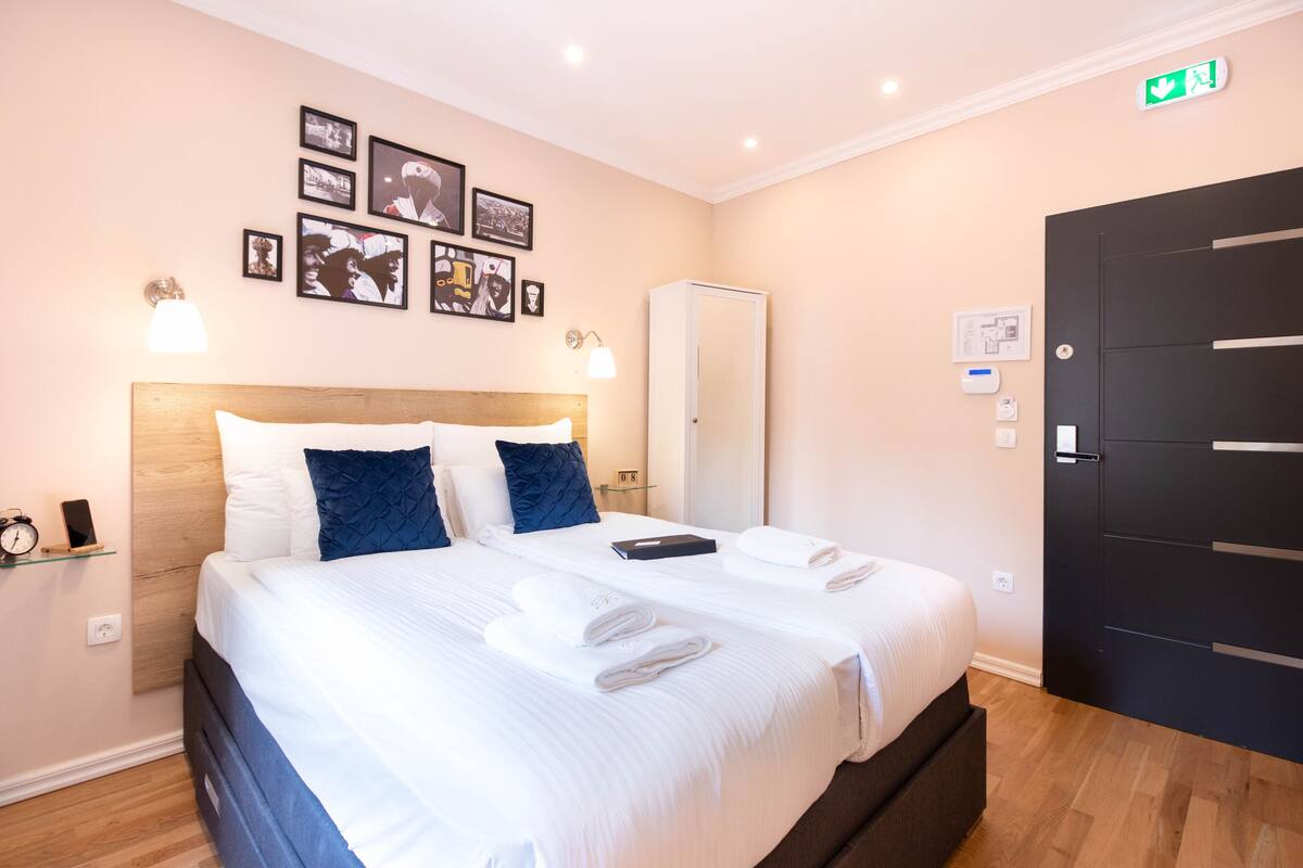 Habitación de hotel con una cama grande, dos almohadas, una mesita de noche con lámpara, un armario y cuadros en la pared.