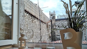 Une vue par la fenêtre sur un mur de pierre, un pot de fleurs, une bougie et un verre d’eau posés sur un comptoir.