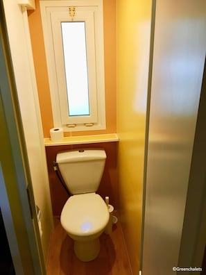 Un baño pequeño y estrecho con un inodoro, una ventana y una repisa con un rollo de papel higiénico.