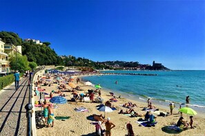 Una playa concurrida con gente tomando el sol y nadando, un cielo azul despejado y edificios en la colina.