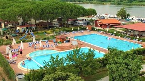 Uma grande piscina ao ar livre com várias pistas de natação, rodeada por espreguiçadeiras e guarda-sóis, com vista para árvores e um lago.