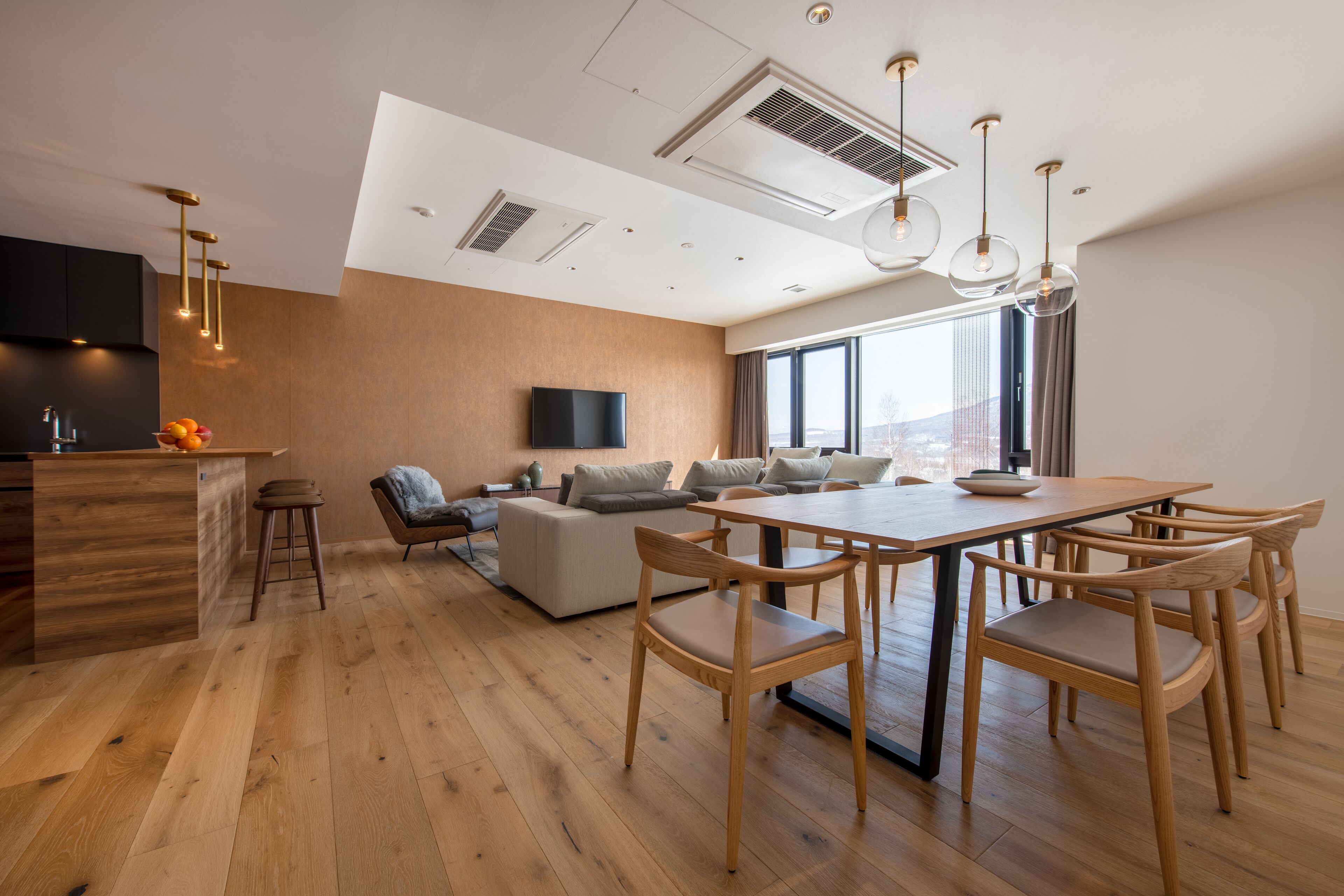 A modern living room with a wooden dining table, comfortable seating, a flat-screen TV, and a large window offering a city view.