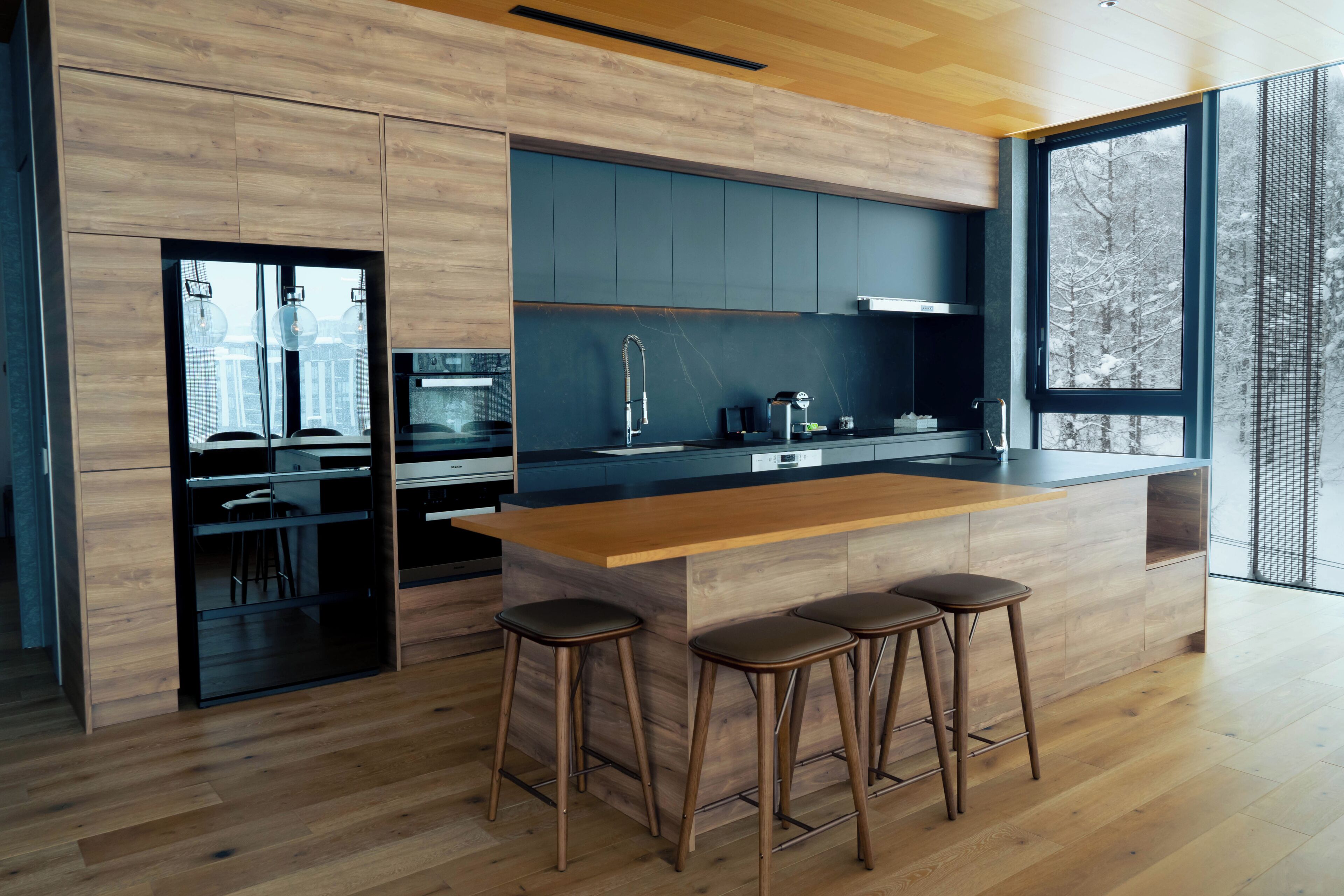 A modern kitchen with wooden cabinetry, a large island with stools, and a view of trees through a window.