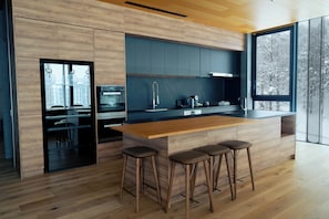 Une cuisine moderne avec des meubles en bois, un grand îlot central doté de tabourets et une vue sur les arbres par la fenêtre.