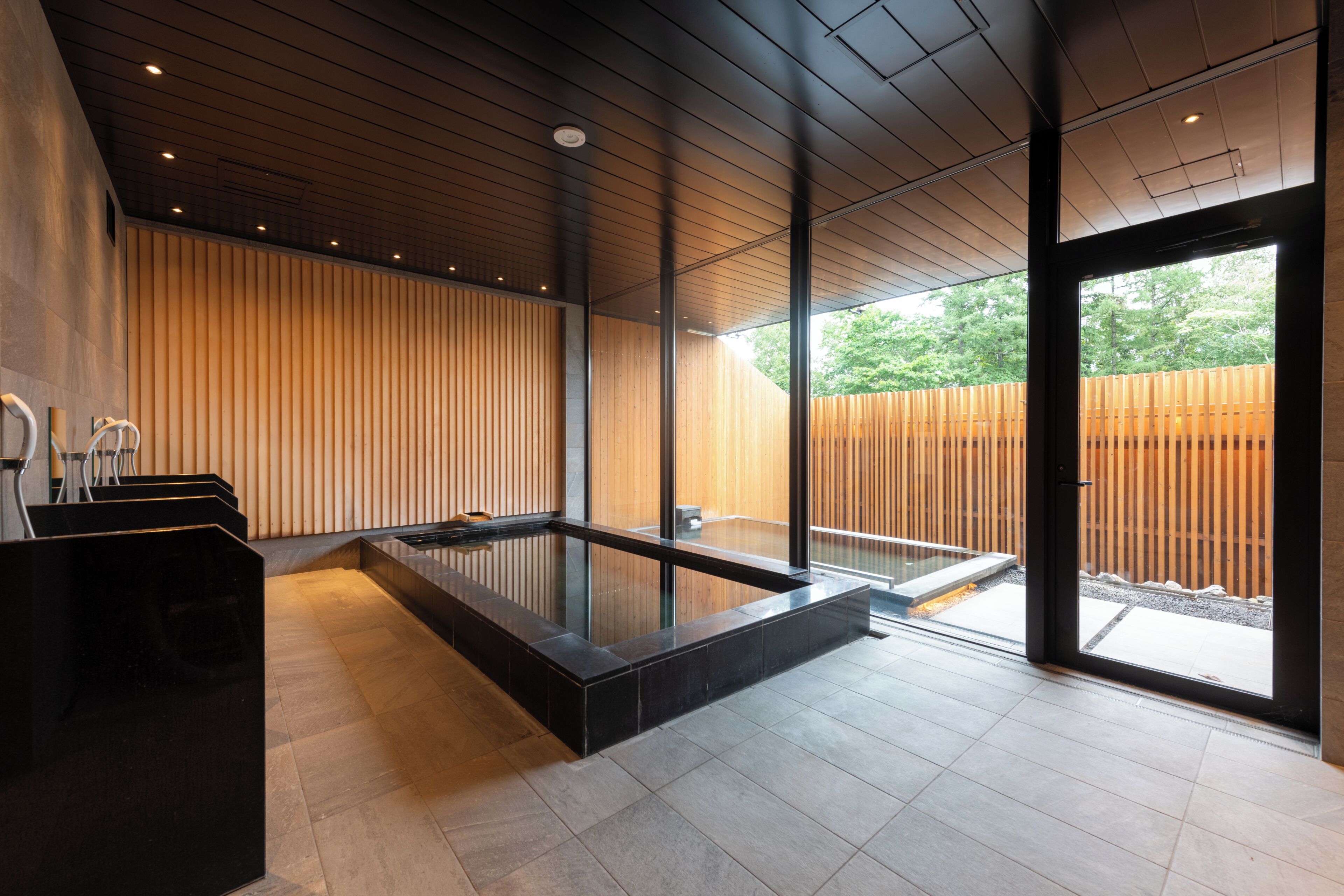 A modern spa area with a wooden wall, a black heated floor, and a glass-enclosed hot tub.