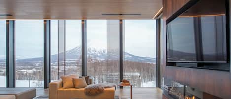 A modern living room with a large TV, a fireplace, and a sofa facing a large window with a mountain view.