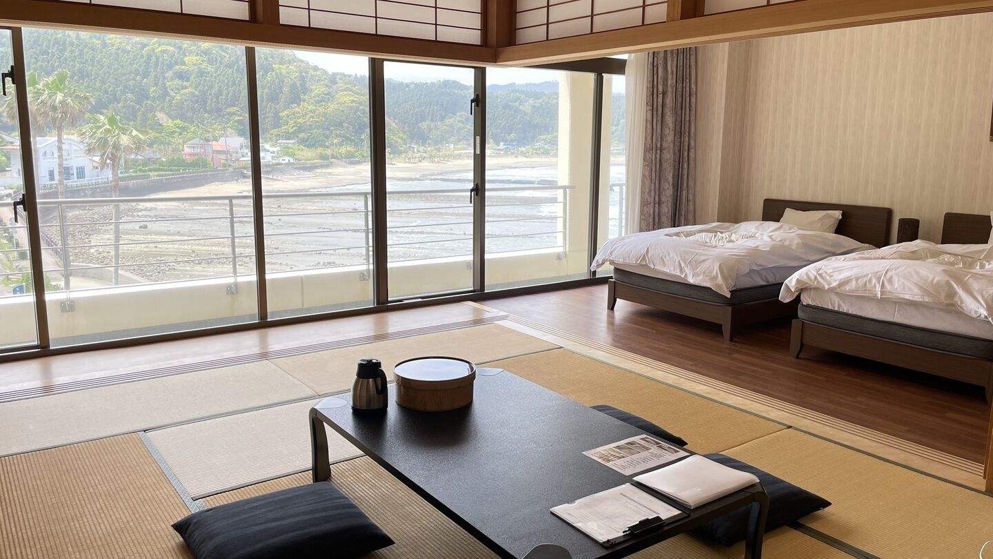 A hotel room with two beds, a tatami mat floor, a table with a kettle and a tray, and a view of a residential area through large windows.