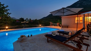 A swimming pool with lounge chairs and a table, surrounded by a stone deck and a mountain backdrop. - Villa Melita with heated pool in Biokovo Nature Park near Makarska (Zagvozd)
