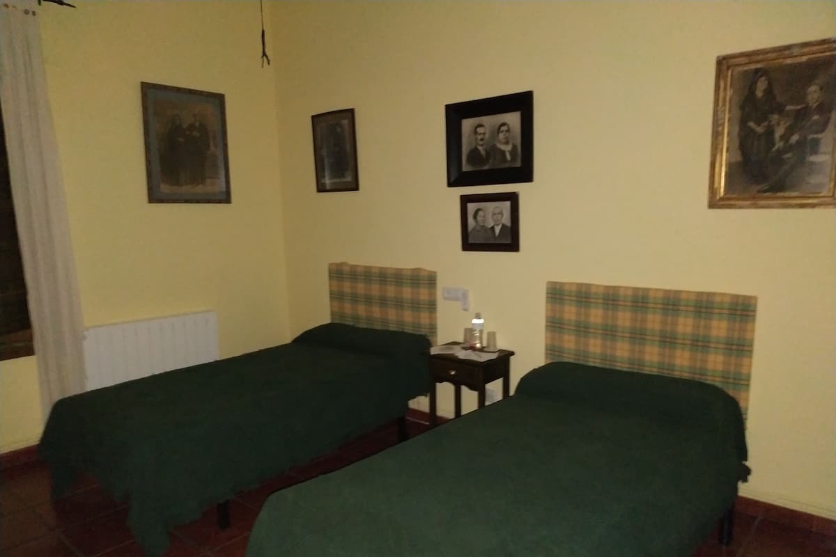 A room with two single beds, a small table with a lamp, and several framed pictures on the wall.