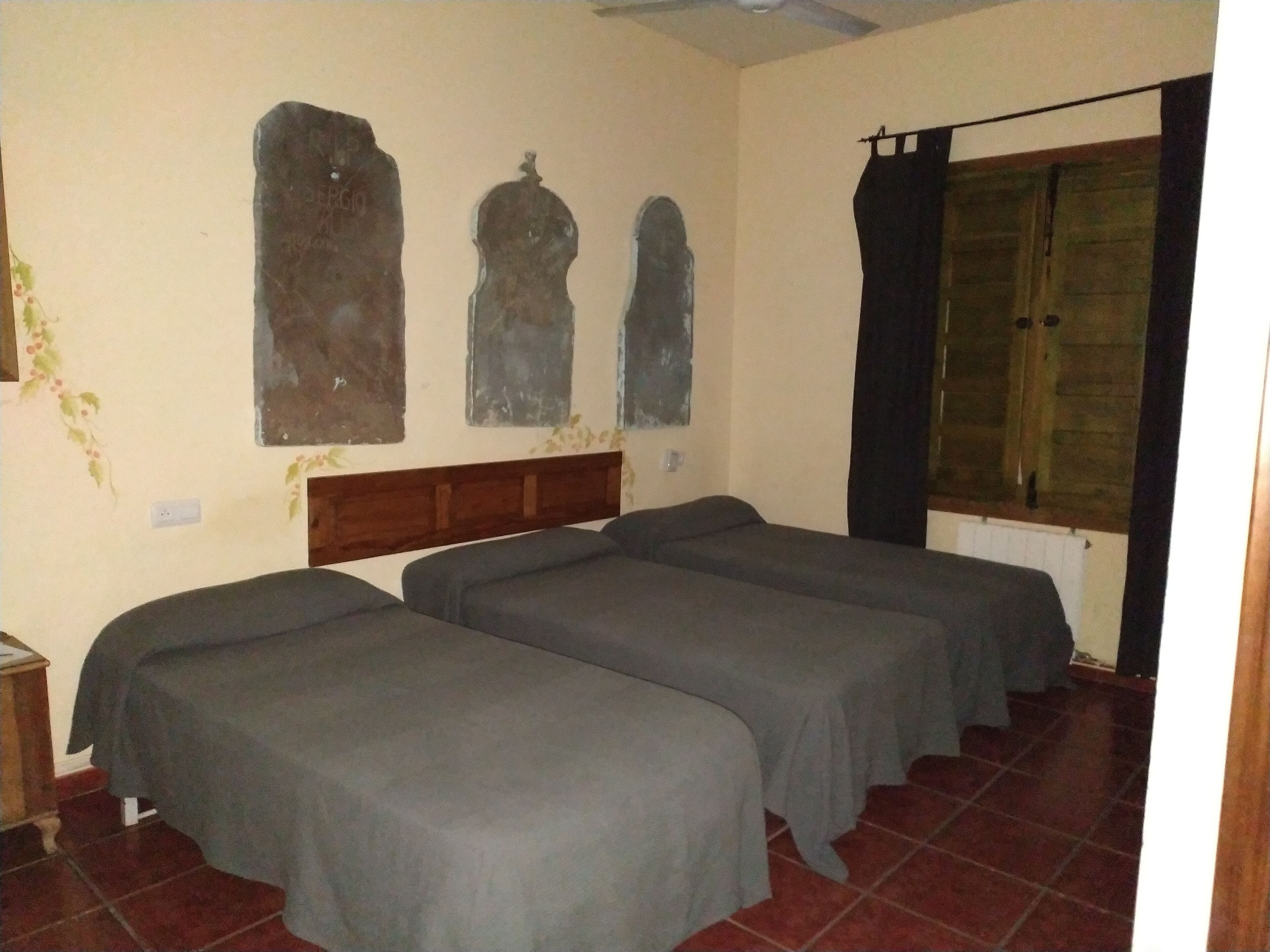 Una habitación con tres camas individuales, cabecera de madera, ventana con cortinas y una obra de arte colgada en la pared.