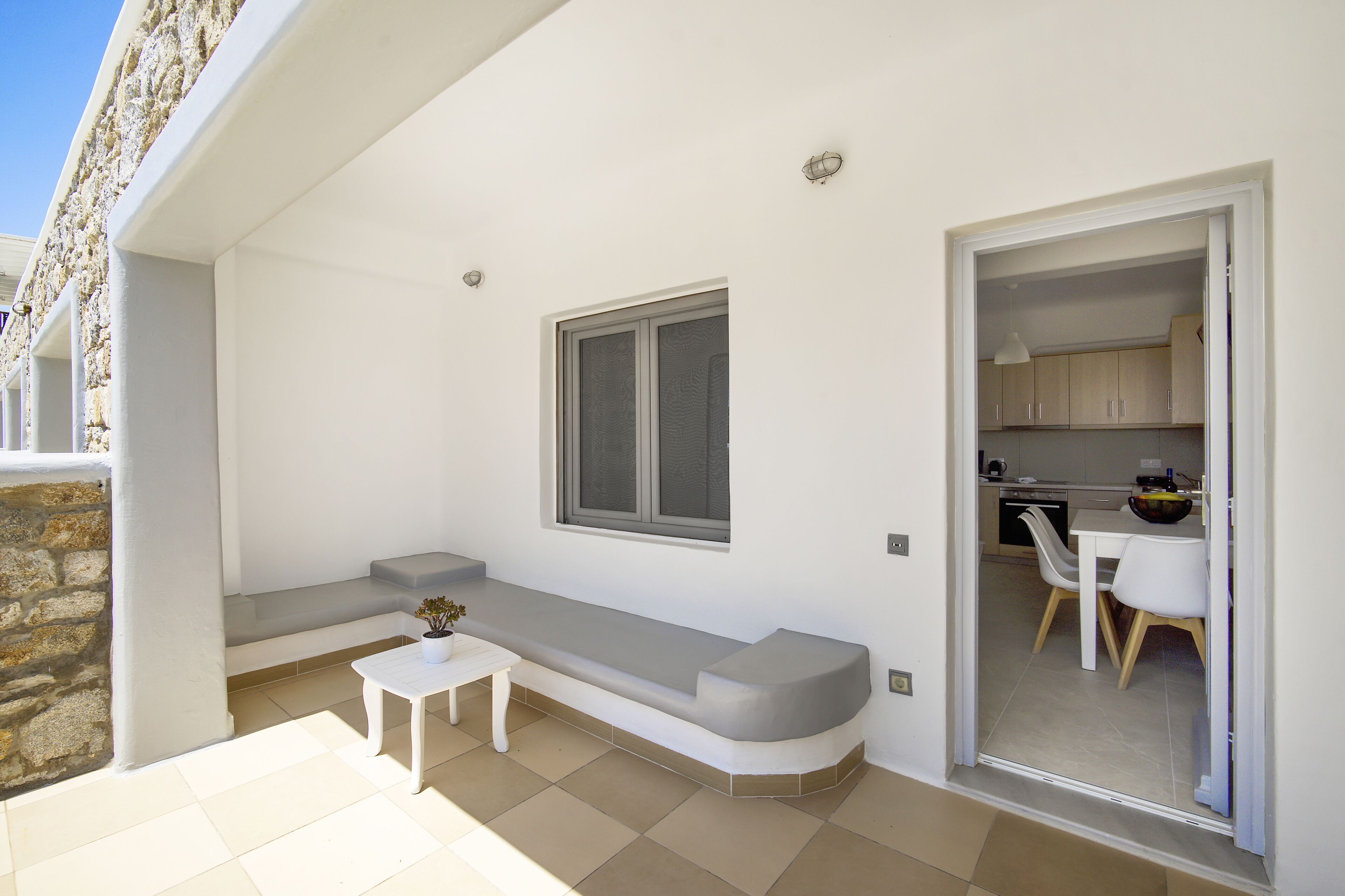 A modern apartment with a balcony, a built-in bench, a small table, and a kitchen area visible through an open door.