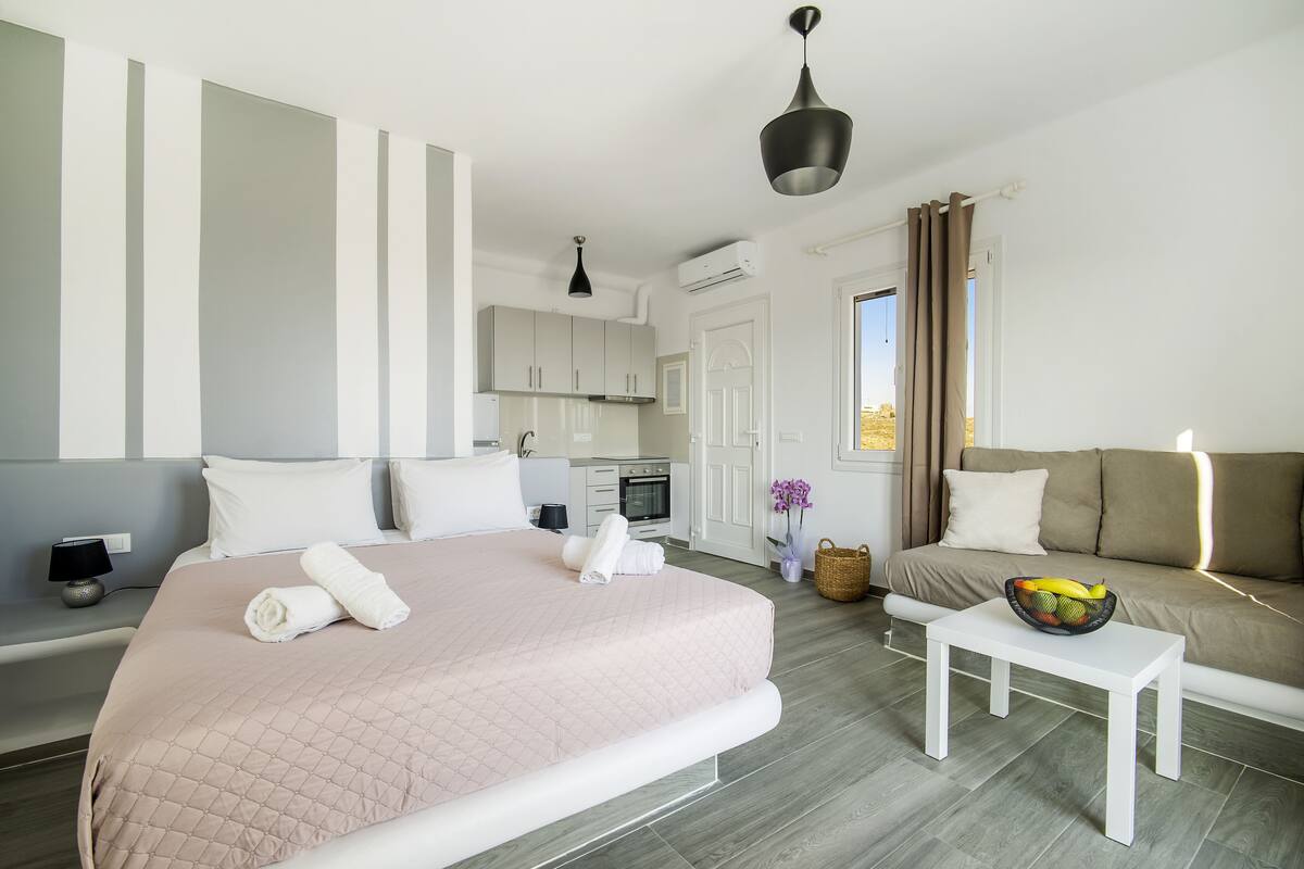 A modern hotel room with a large bed, a sofa, a small table, and a kitchen area in the background.