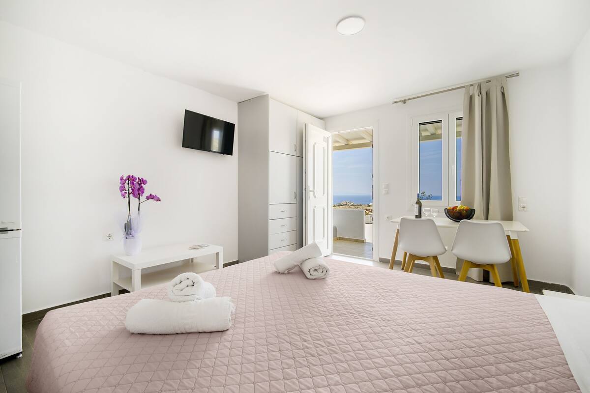 A hotel room with a bed, a desk, a chair, a TV, and a view of the sea.