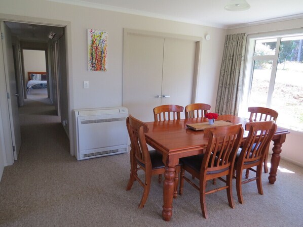 A dining area with a wooden table and chairs, a white air conditioner, and a view of the outdoors through a large window.