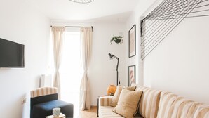 A living room with a striped sofa, a chair, a coffee table with books, a TV, and a window with curtains. - Nephiria's Boho House with Acropolis View (Athens)