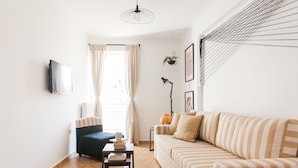 A living room with a sofa, a chair, a coffee table with books, a TV mounted on the wall, and a window with curtains. - Nephiria's Boho House with Acropolis View (Athens)