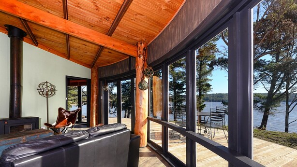 Un interior moderno con techo de madera, ventanas amplias, un sofá de cuero negro y vistas a un lago.
