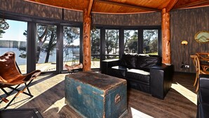 Interior acogedor de una cabaña con techo de madera, ventanas amplias, un sofá de cuero negro, un cofre azul y una silla de madera.