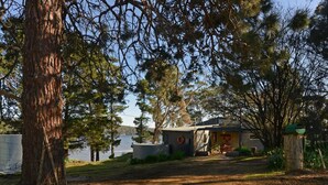 Una cabaña rústica con un camino de tierra, rodeada de árboles altos y un lago.
