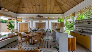 Een ruime kamer met hoge plafonds, een eethoek met tafel en stoelen, een keuken met een aanrecht en spoelbak, en uitzicht op groen buiten.