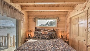 A cozy wooden cabin bedroom with a large bed, patterned bedding, a window with a view, and a wooden door. - Rio Grande National Forest Cabin: Views & Hot Tub (Del Norte)