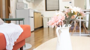 A room with a wooden floor, a white vase with flowers on a round table, a red sofa with a white blanket, and a kitchen in the background. - Elle’s Shirokane guest house (Tokyo)