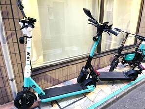 Two electric scooters parked on a tiled pavement in front of a building with a window. - Elle’s Shirokane guest house (Tokyo)
