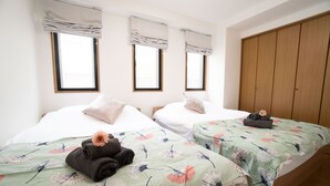 A hotel room with two single beds, each with a floral bedspread and a window with a valance. There is a wooden wardrobe and a small table with towels and a flower. - Elle’s Shirokane guest house (Tokyo)