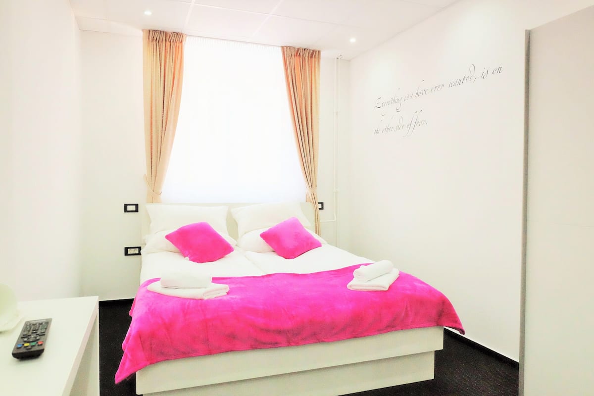 A hotel room with a bed, pillows, a blanket, a remote control, and a wall with a quote.