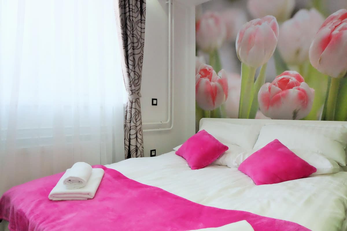 A neatly made bed with white and pink bedding, a rolled towel, and a wall with a tulip painting.