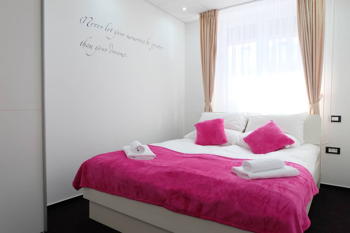 A neatly made bed with a pink blanket and pillows, a white wall with a motivational quote, and a window with sheer curtains.