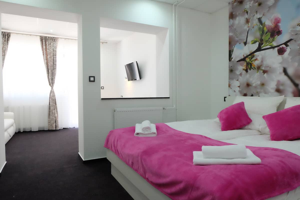 A hotel room with a bed, a white chair, a mirror, and a wall with a floral painting.