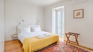 A bedroom with a bed, bedside lamps, a yellow bedspread, a patterned rug, a wooden drying rack, and a framed picture on the wall. - Chiado 1 - Carmo Square - Recently renovated apartment with great view of the River Taugus and São J (Lisbon)