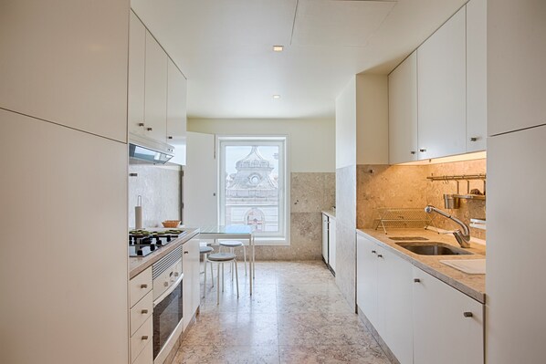 A narrow kitchen with white cabinets, a marble countertop, a sink, and a window with a view of a building.