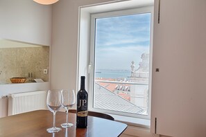 A room with a view of a cityscape through a window. There is a bottle of wine and two glasses on a wooden table.