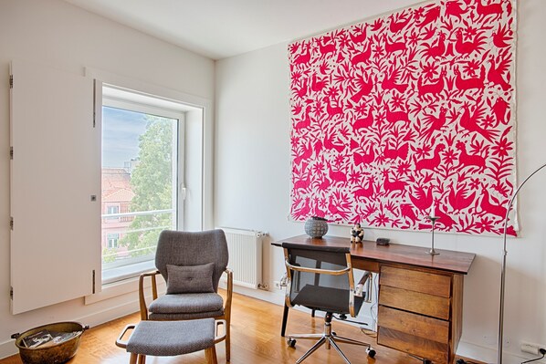 A room with a large window, a chair, a desk, and a red patterned tapestry.